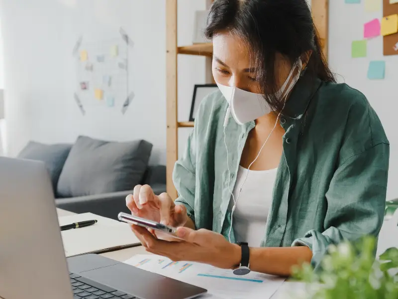 Freelancer Asia women wear face mask using smartphone shopping online via website while sitting at desk in living room.